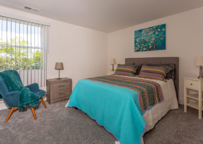 Furnished bedroom with a queen bed, teal accent chair, carpet flooring, and large windows with blinds at Creekside Apartments in Bensalem